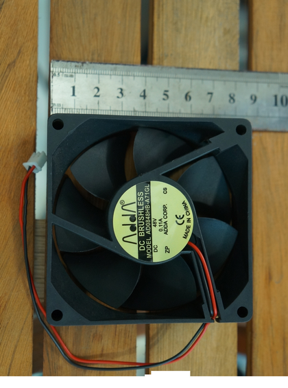 External reference: ADDA AD0848HB-A71GL, DC48V 0.11A; two red/black leads, wire loop and a pale two-position connector housing. Background edited. Different unit; ruler does not establish supplied dimensions. Actual condition, full option, connector and pinout require verification.